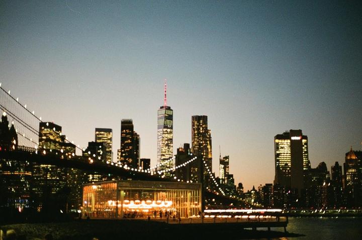A carousel and the Brooklyn Bridge are visible in front of the illuminated New York City skyline at dusk.