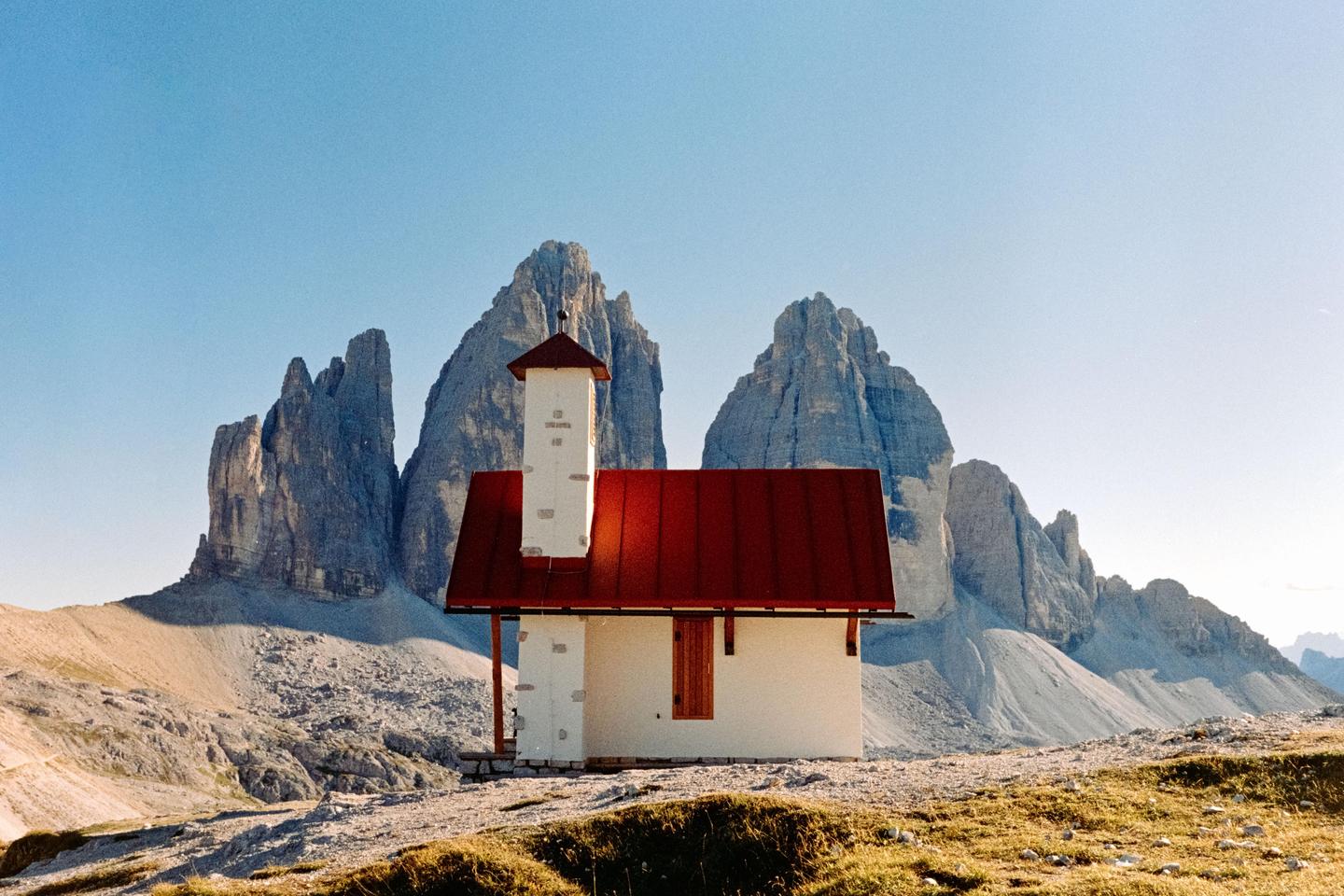 A small white church with a red roof stands in a rocky, mountainous landscape under a clear sky.