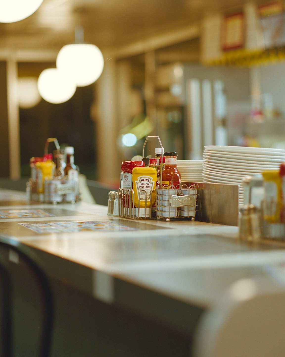 Condiments and stacked plates sit on a diner counter under glowing spherical lights.
