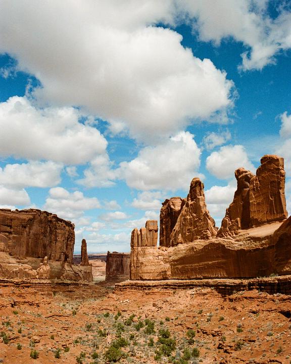 A vast desert landscape features towering red rock formations under a bright blue sky with fluffy white clouds.