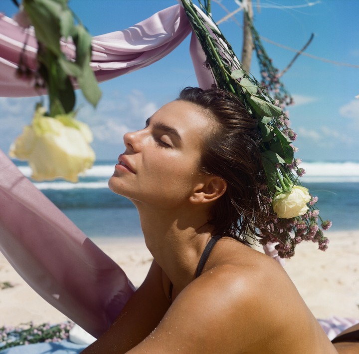 A woman with closed eyes and wet hair wears a flower crown on a beach.