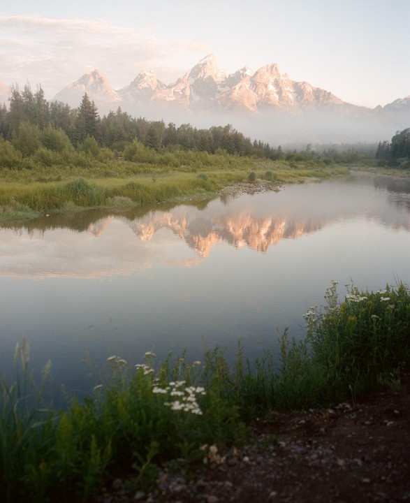 A river reflects mountains and trees under a hazy sky.