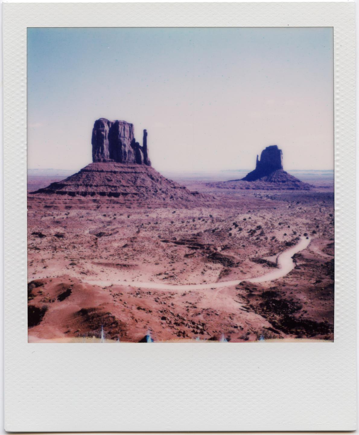 Two large rock formations stand in a vast desert landscape with a winding dirt road.