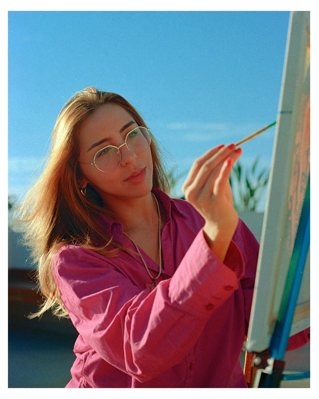 A woman wearing glasses and a pink shirt is painting on a canvas under a clear blue sky.