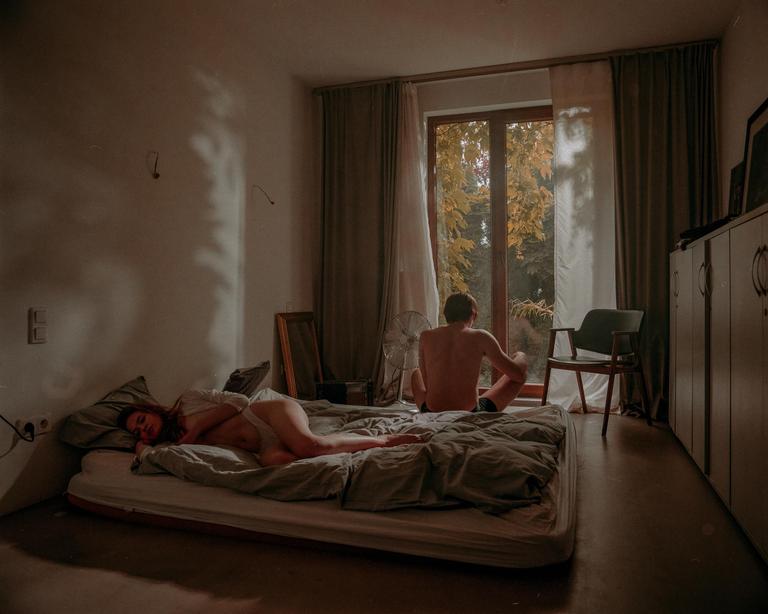 A woman lies on a mattress on the floor while a man sits on the floor looking out a window at autumn foliage.