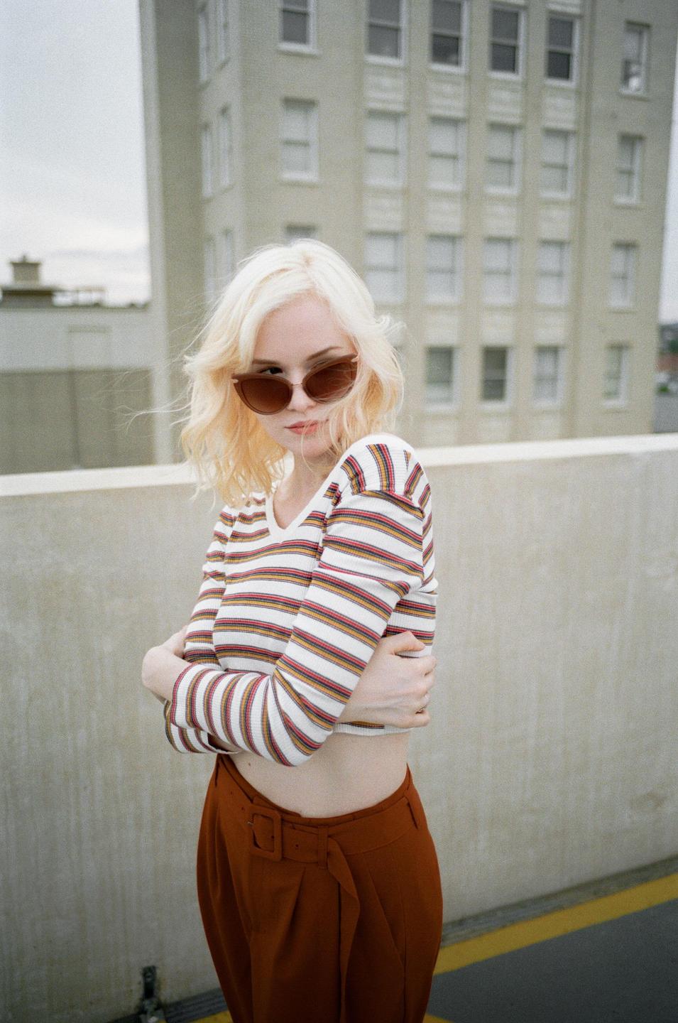A woman with blonde hair and sunglasses stands with her arms crossed in front of a building.