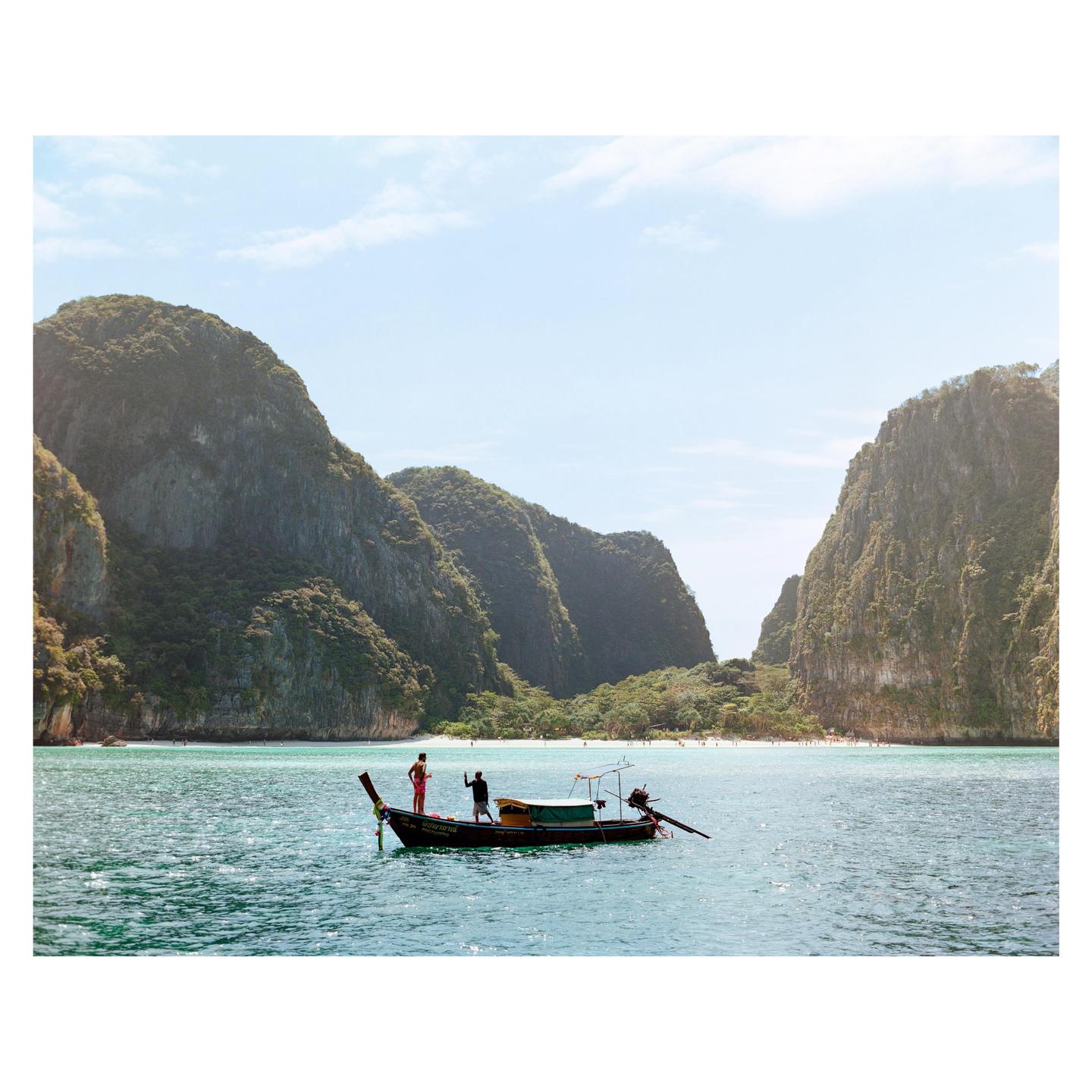 Two men stand on a longtail boat in clear blue water with large, green cliffs and a sandy beach in the background.