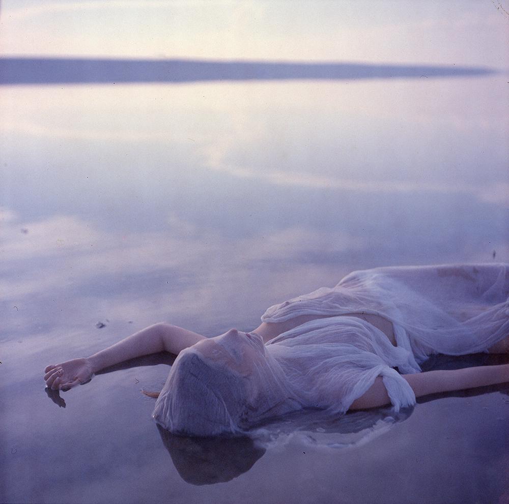 A woman in a white dress lies in shallow water with her face partially obscured by fabric.