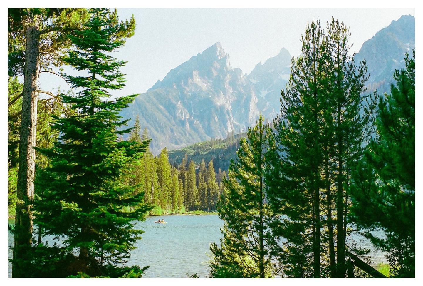 A lake with two people in a boat is surrounded by a forest with mountains in the background.