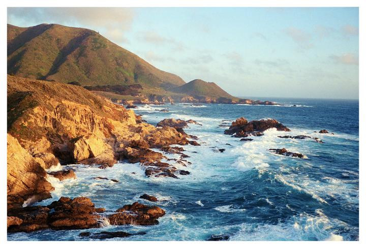 Waves crash against a rocky coastline with green hills in the background under a light blue sky.