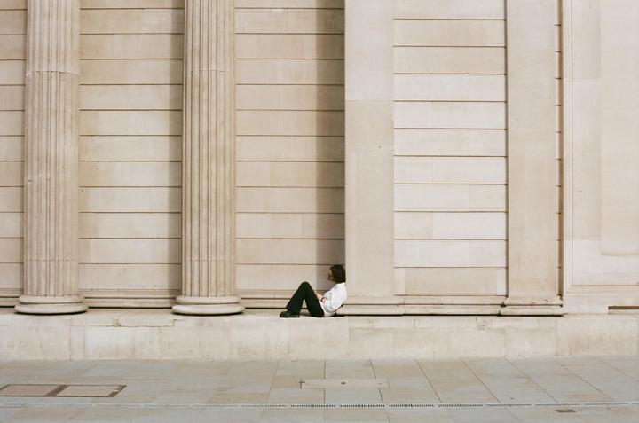 A man sits on a ledge in front of a large stone building with columns.