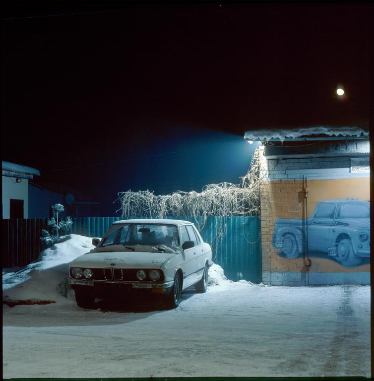 A white car is parked in a snowy yard next to a garage with a truck painted on its door at night.