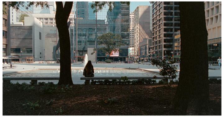 A woman sits on a bench in front of a fountain in a city plaza with tall buildings in the background.