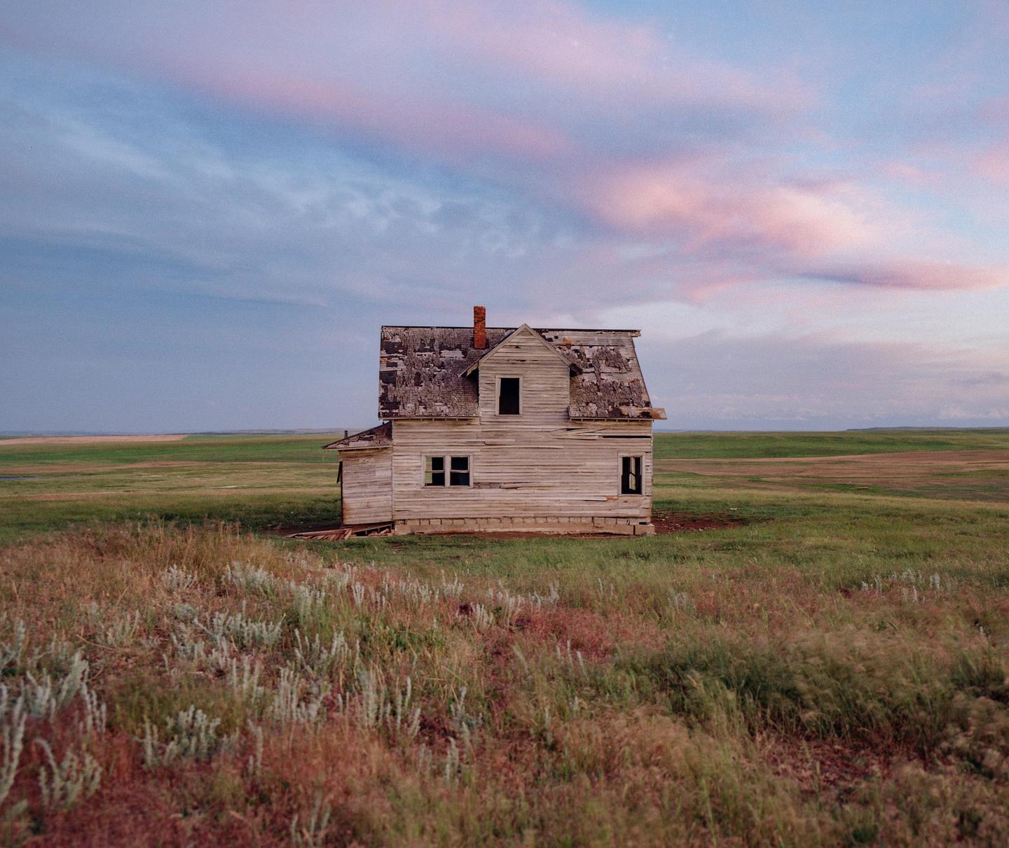 An abandoned house stands in a vast field under a sky with pink clouds.