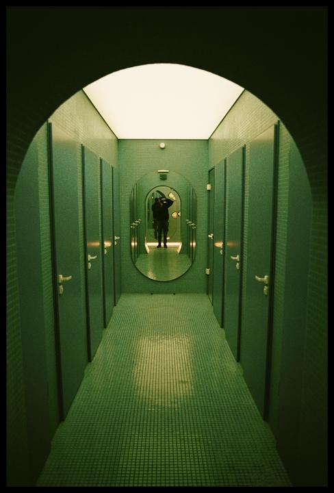 A person is reflected in an oval mirror at the end of a long, tiled hallway with stalls on either side.