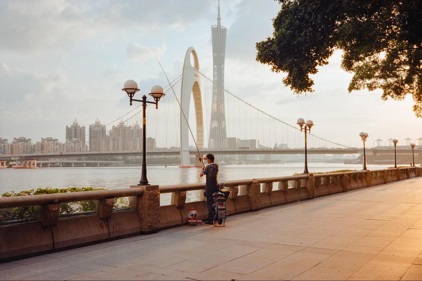 A man fishes from a riverbank promenade with a city skyline and bridge in the background at sunset.