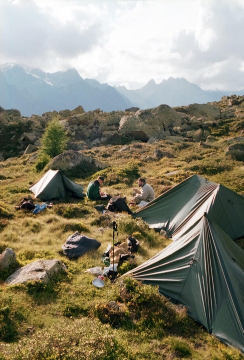 Two men sit on a grassy, rocky mountain slope next to tents, with mountains in the background.