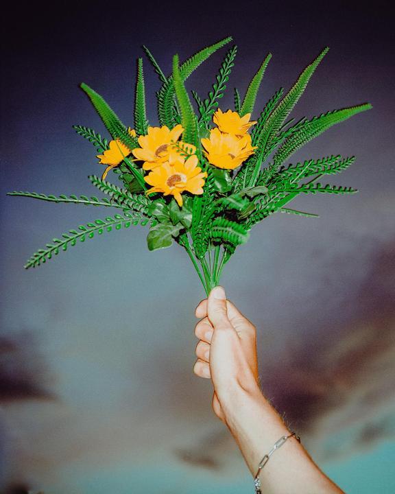 A hand holds a bouquet of artificial yellow flowers and green foliage against a cloudy sky.