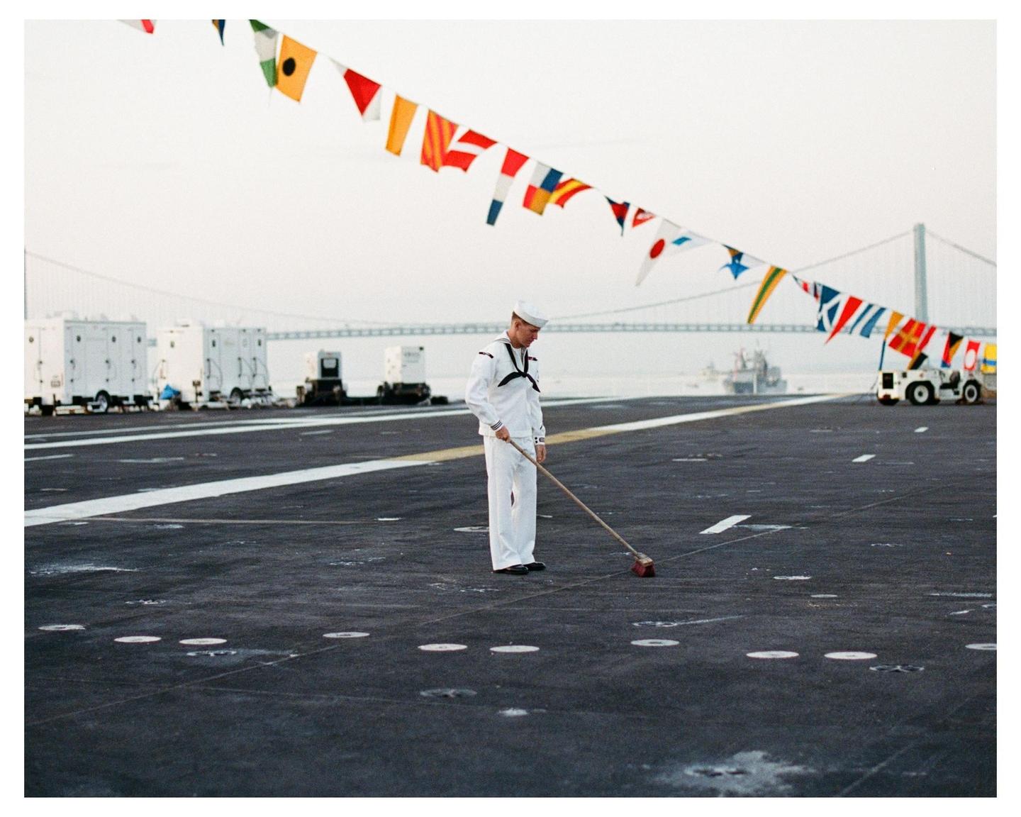 A man in a white uniform sweeps the deck of a ship adorned with colorful signal flags.