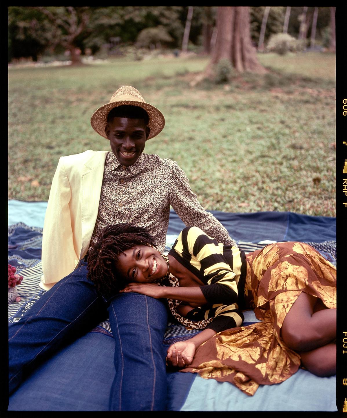 A man in a straw hat sits on a blanket with a woman resting her head on his lap in a grassy area.