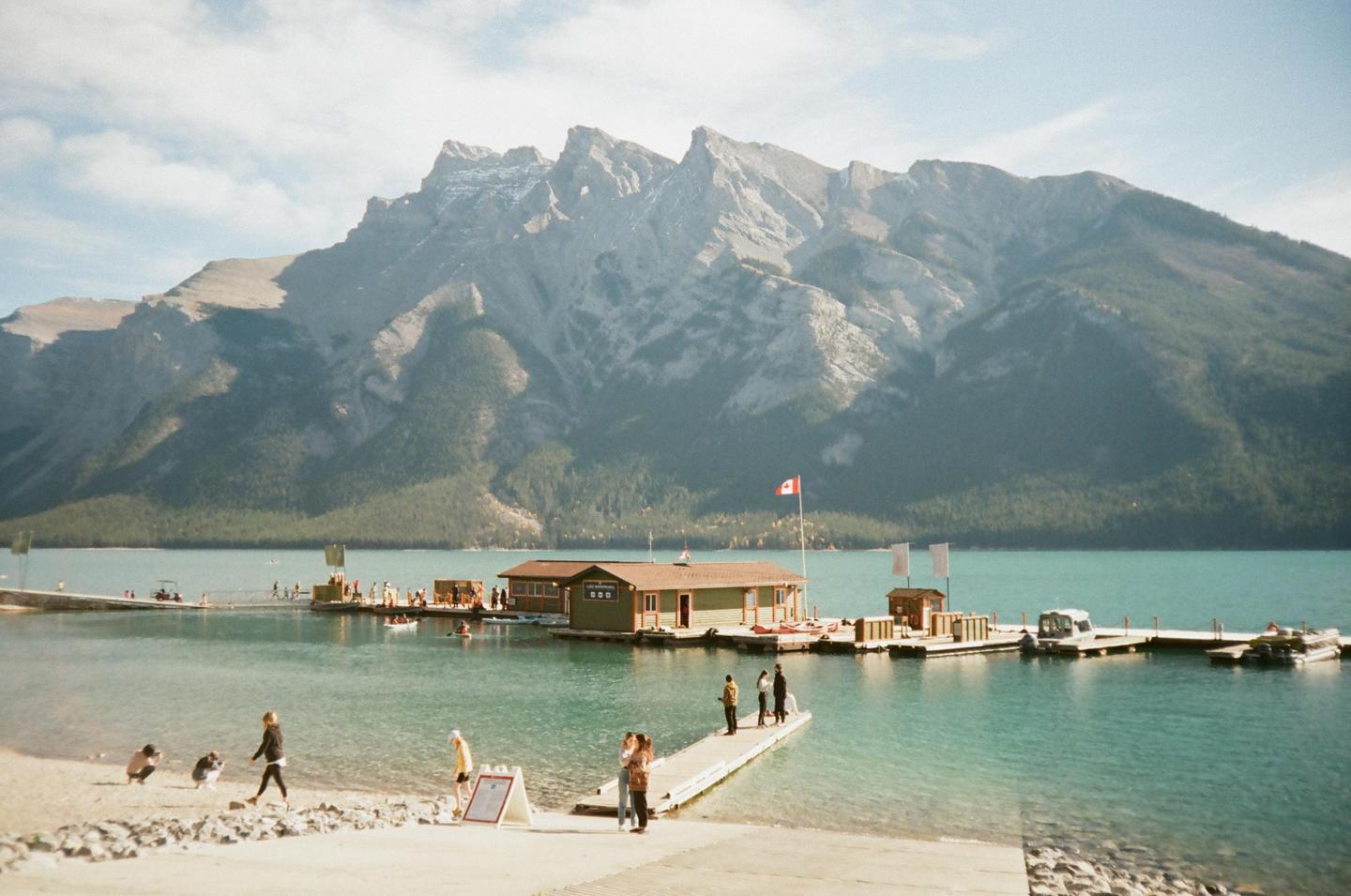 A Canadian flag flies over a boathouse on a turquoise lake with mountains in the background.