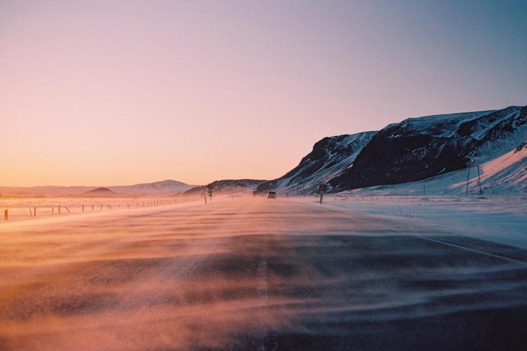 A car drives on a snowy road with mountains in the background under a colorful sky.