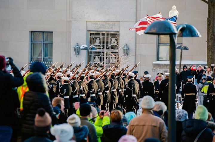A line of soldiers in uniform march with rifles past a building with an American flag flying above it, as a crowd of people watch.