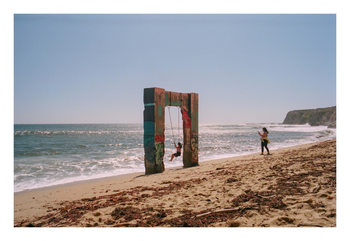 A woman swings on a swing set made from concrete pillars on a beach while another woman takes a photo.