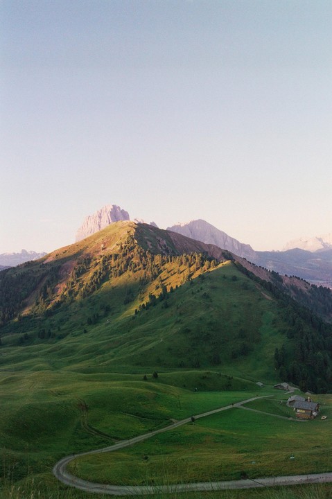 A winding road leads to a small cluster of buildings nestled in a green valley with mountains in the background.