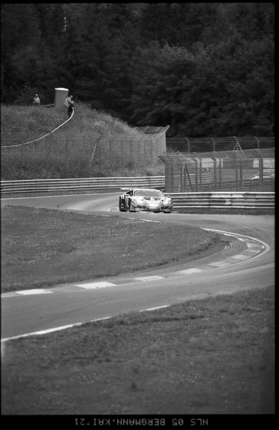 A race car speeds around a turn on a track with two men watching from a grassy hill.