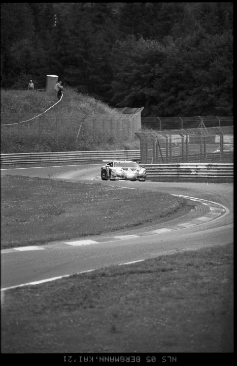 A race car speeds around a turn on a track with two men watching from a grassy hill.