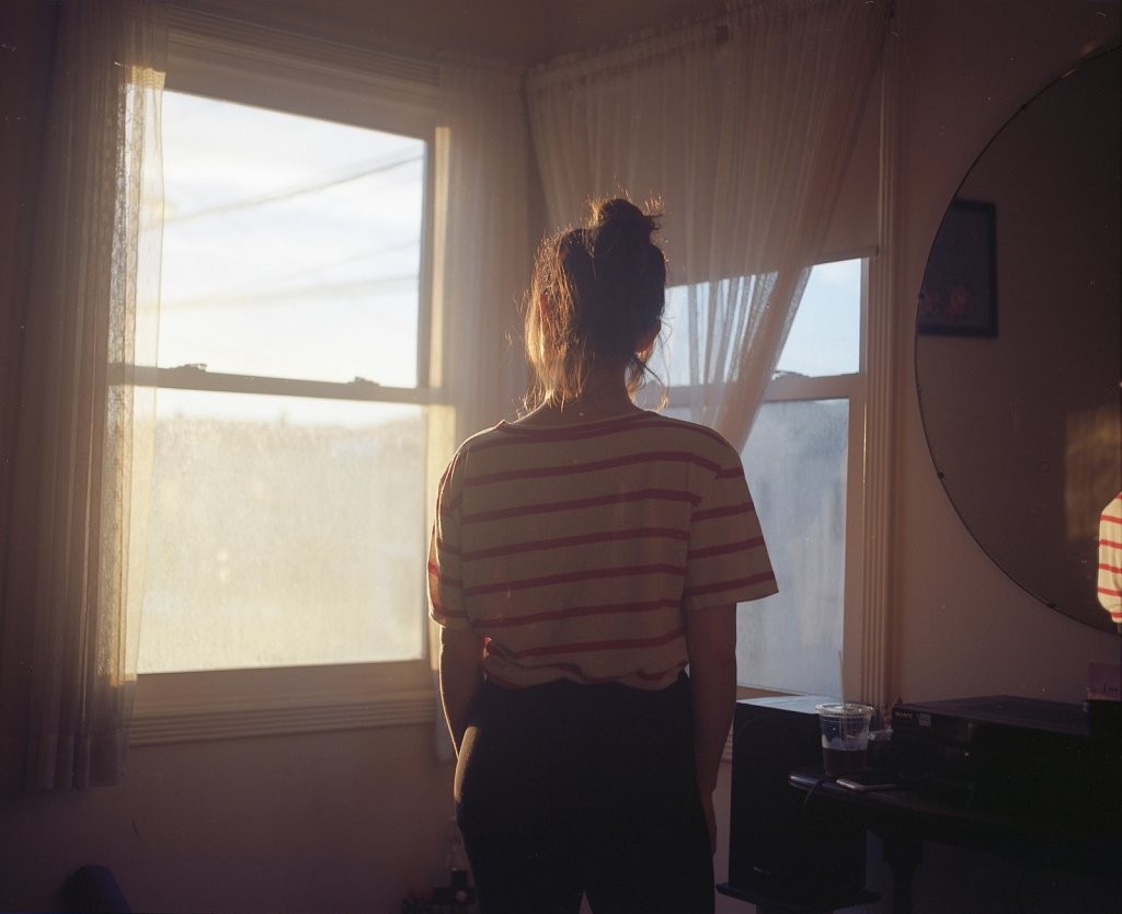 A woman stands with her back to the camera, looking out a window at sunset.