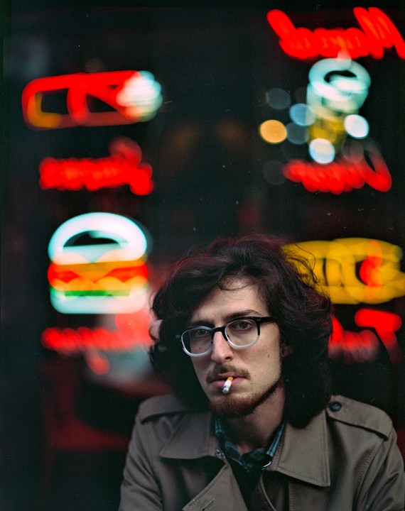A man with glasses and a beard smokes a cigarette in front of blurred neon signs at night.