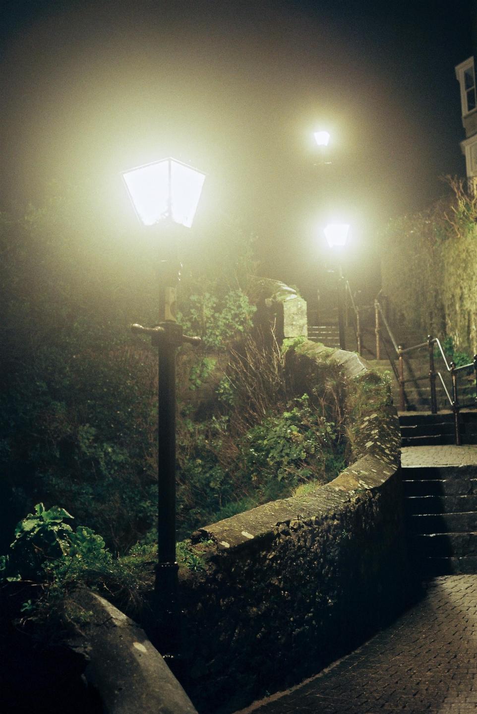 A foggy night scene shows a stone wall and steps illuminated by three streetlights.