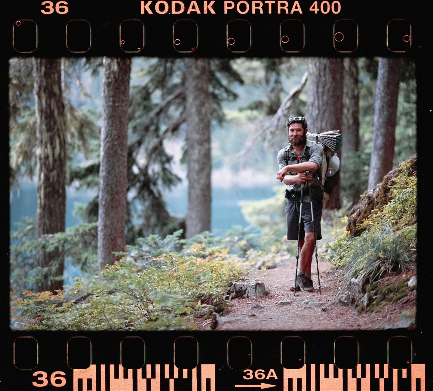 A man with a beard and backpack stands on a trail in a forest next to a body of water.