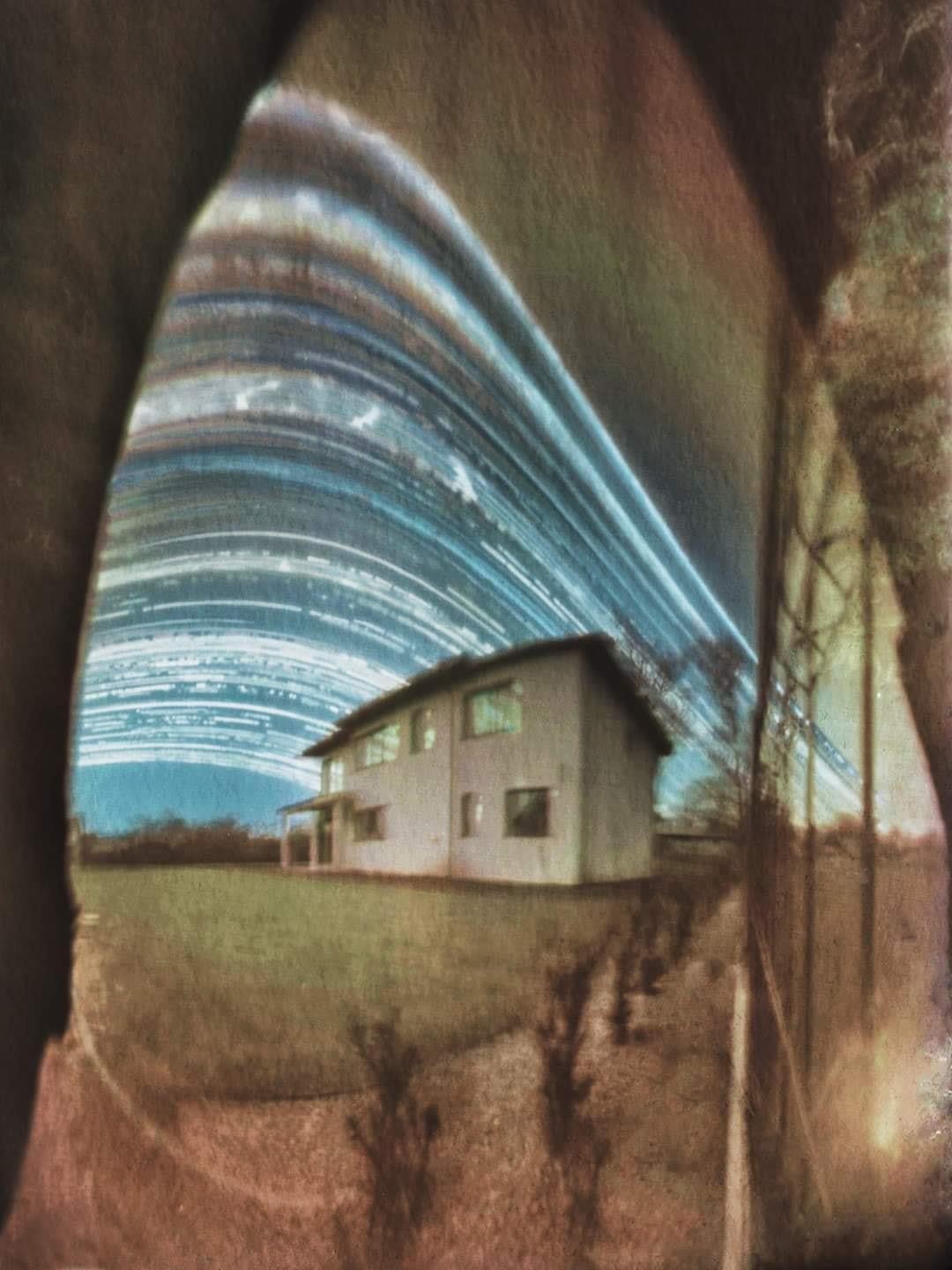 A house stands in a field under a sky with long star trails.