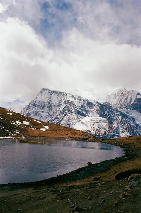 A large body of water sits in the foreground with snow-capped mountains and a cloudy sky in the background.