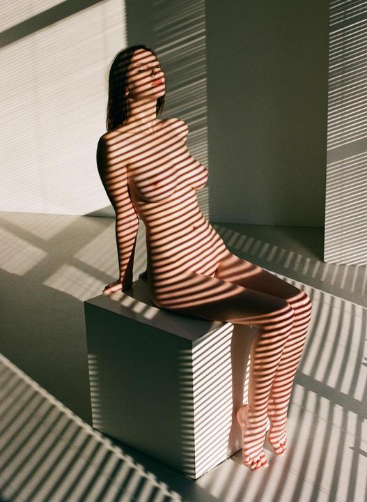 A woman sits nude on a white block with shadows from blinds cast across her body and the floor.