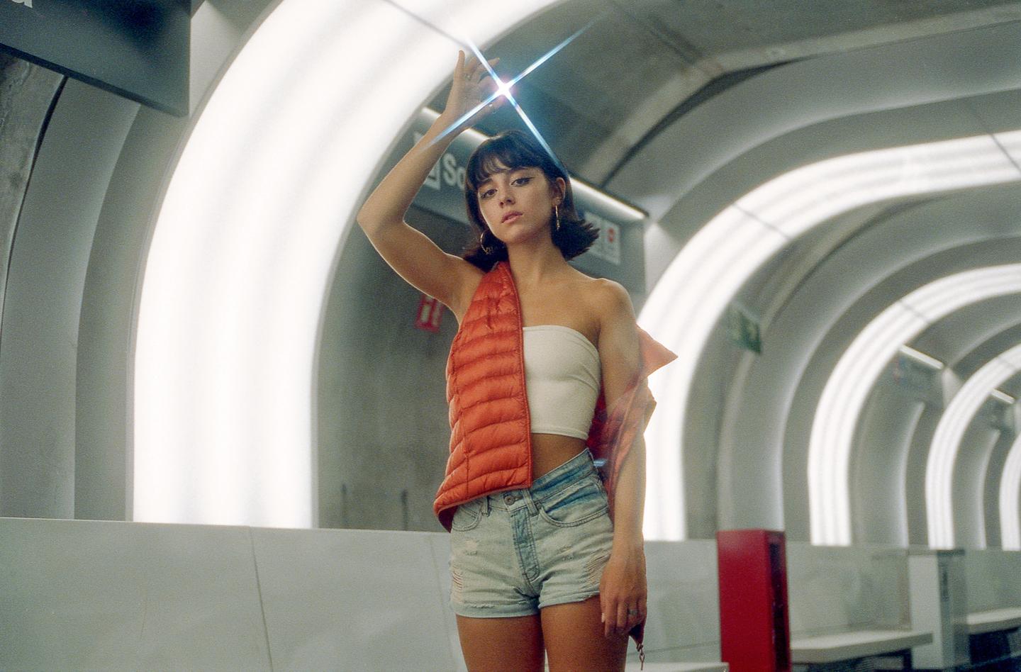 A woman in a white tube top, denim shorts, and an orange vest stands in a subway station with her arm raised, catching a starburst flare from a light.