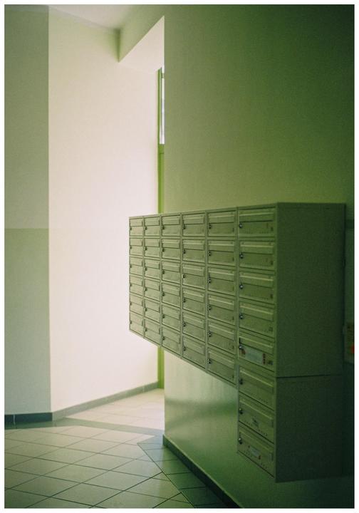 A wall of mailboxes is mounted on a green wall in a hallway with a tiled floor.