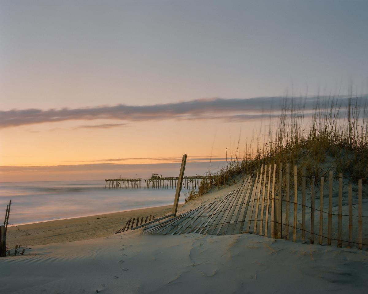A wooden fence and dune grass stand on a sandy beach with a pier in the distance at sunset.