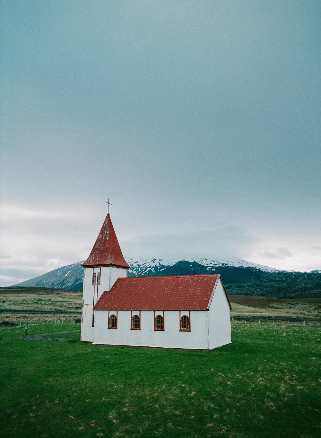 A white church with a red roof stands in a green field with a snow-capped mountain in the background under a cloudy sky.