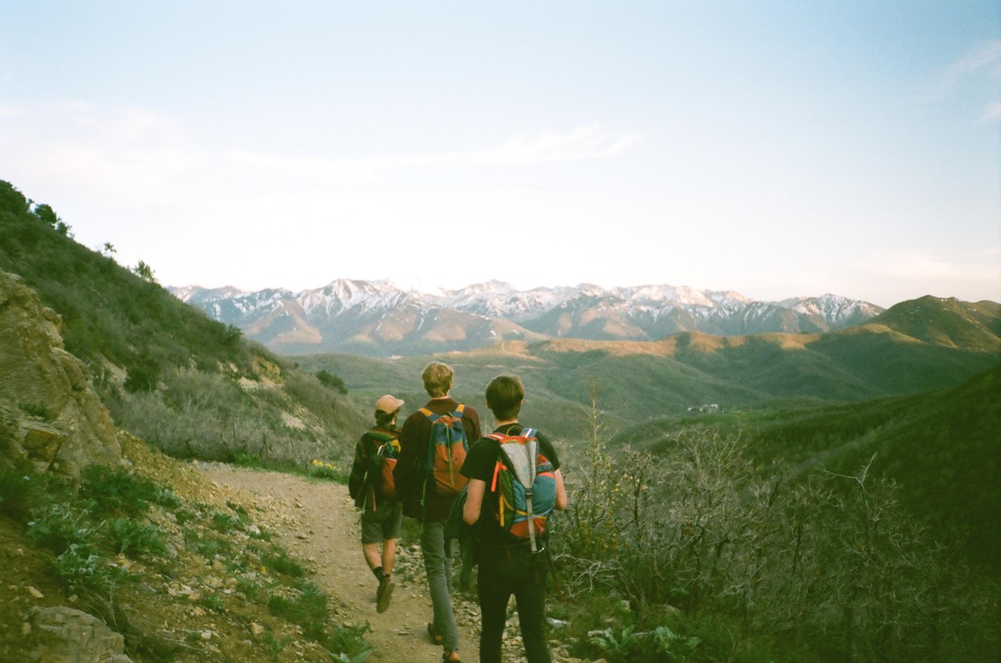 Three people with backpacks walk on a dirt path overlooking a valley and snow-capped mountains.