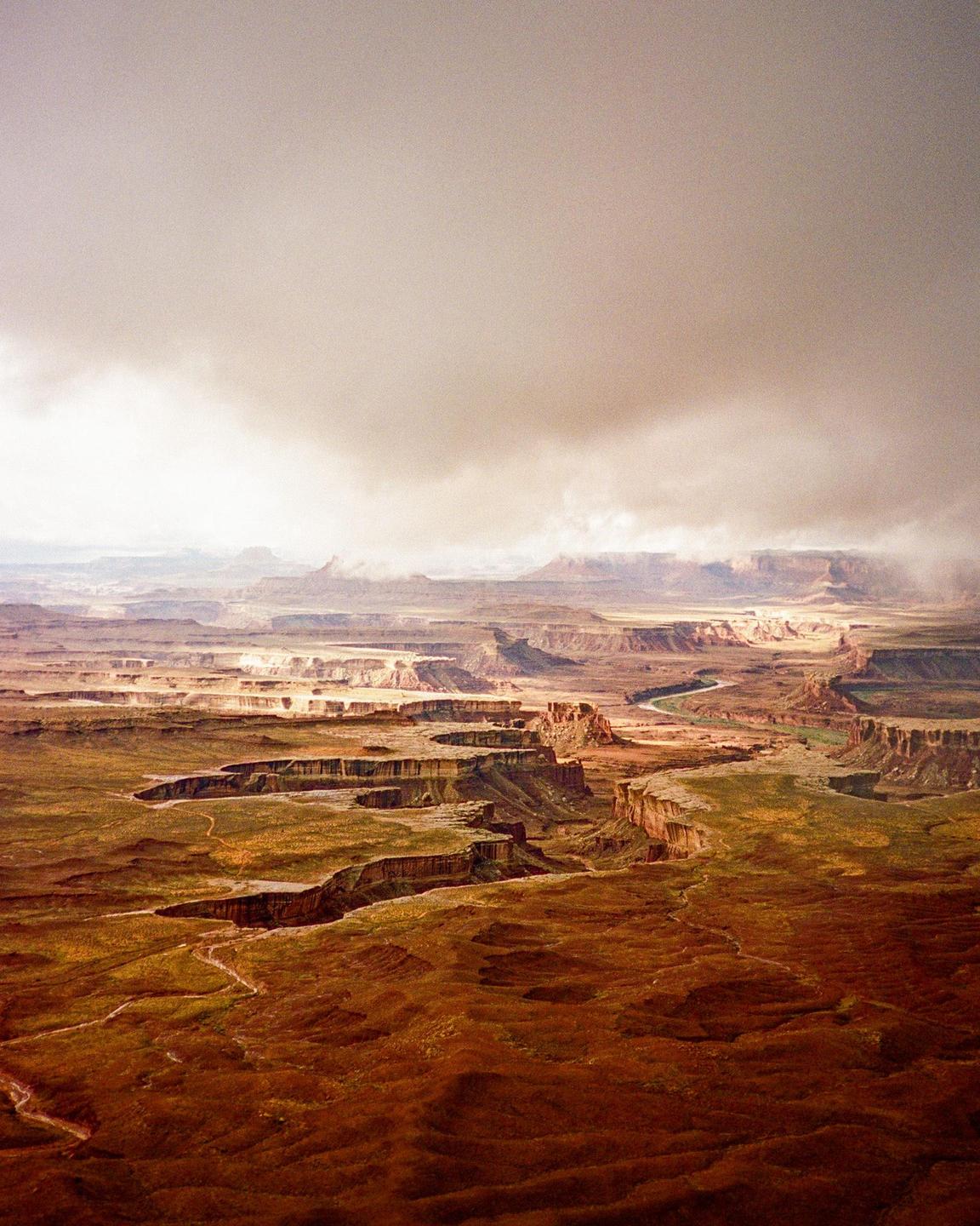 A vast canyon landscape with a river winding through it is visible under a cloudy sky.