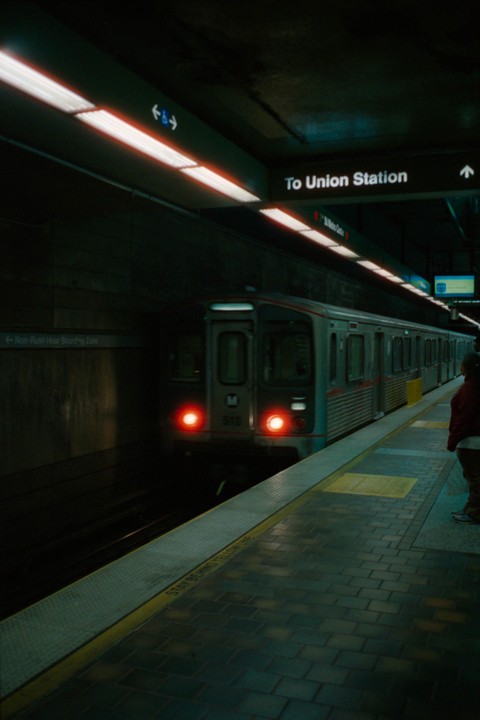 A subway train with red lights on its front is stopped at a dark underground station platform where a person is waiting.