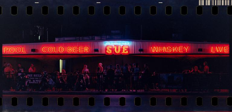 A bar with neon signs for "Pool", "Cold Beer", "Sue", "Whiskey", and "Live" is illuminated at night, with people gathered below.