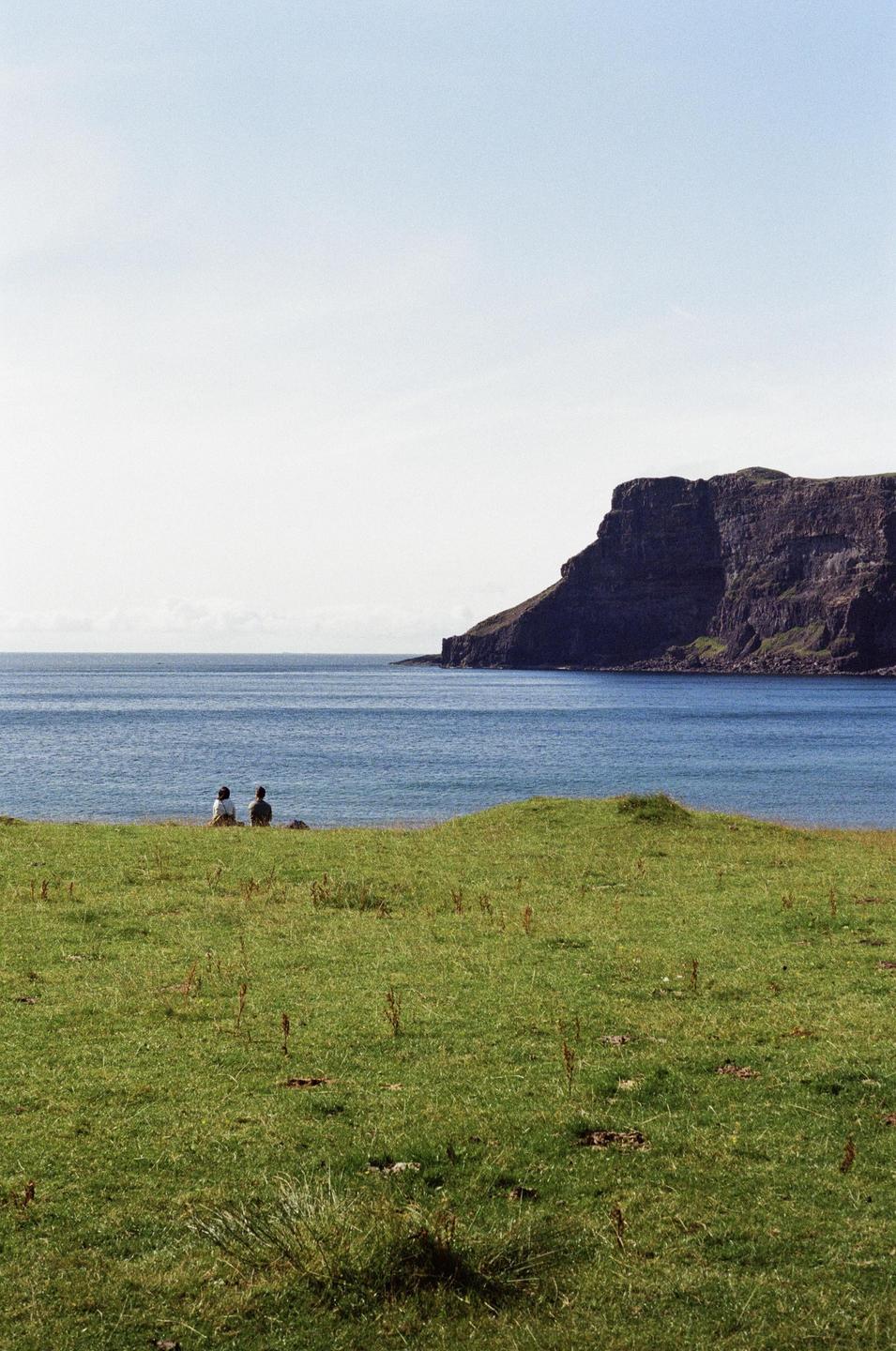 Two people sit on a grassy hill overlooking a body of water and a large cliff.