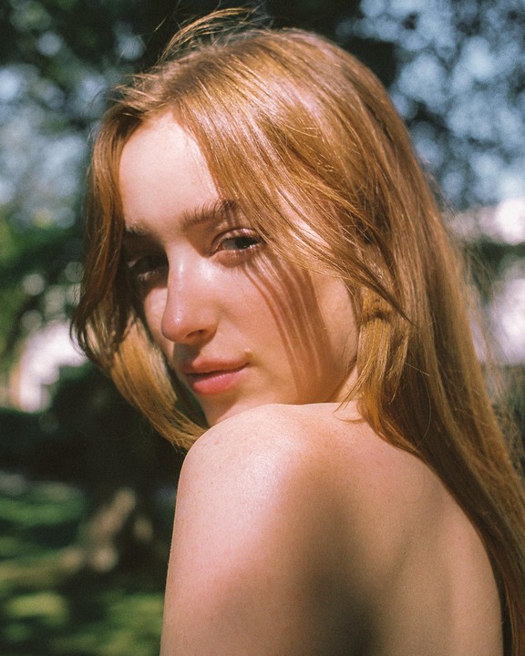 A woman with long red hair looks over her shoulder at the viewer, with shadows cast across her face and shoulder.