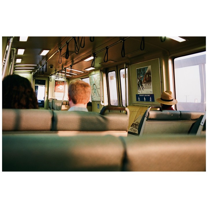 Three people are riding a train, with two seen from behind in the foreground and one further back wearing a hat.