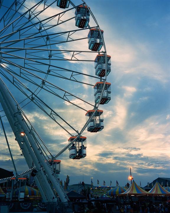 A ferris wheel with illuminated gondolas stands against a cloudy sky at dusk, with carnival tents and people visible below.